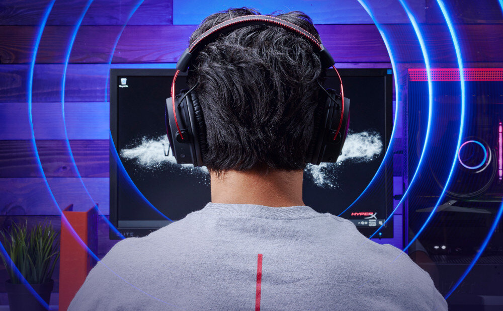 Osoba nosi nauszne słuchawki przed monitorem, na którym widoczny jest napis \'HYPERX\'. W tle znajdują się kolorowe światła i drewniana ściana.
