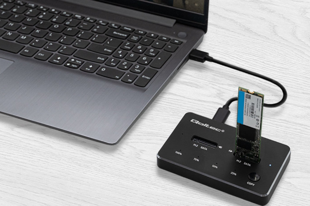 Na białym stole znajduje się laptop podłączony do stacji dokującej Qoltec, w której umieszczony jest dysk SSD. Na stacji widoczne są napisy: \'Qoltec\', \'M.2 SATA\', \'M.2 SATA/IR\', \'100%\', \'75%\', \'50%\', \'25%\', \'0%\', oraz przycisk \'COPY\'.