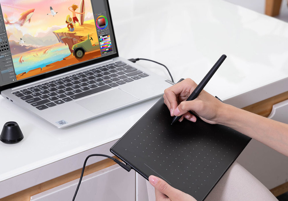 Osoba używa czarnego tabletu graficznego z rysikiem, podłączonego do laptopa, na którym wyświetlany jest kolorowy obrazek. Na biurku znajduje się również podstawka na rysik.