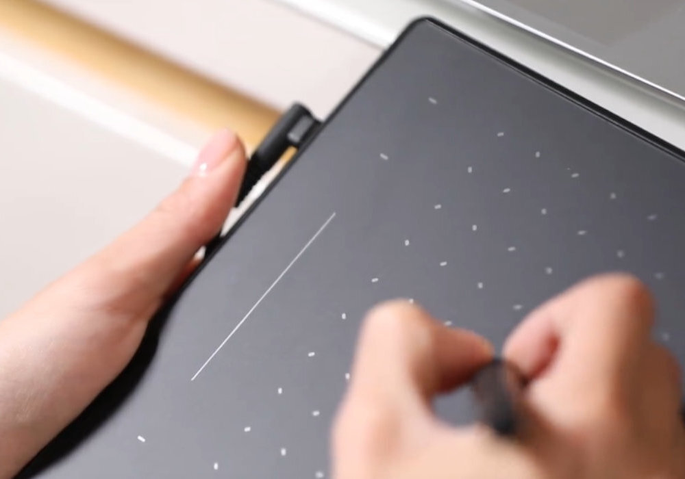 Osoba trzyma czarny tablet graficzny z białymi kropkami i rysuje na nim za pomocą pióra. Tablet jest podłączony do urządzenia za pomocą kabla.