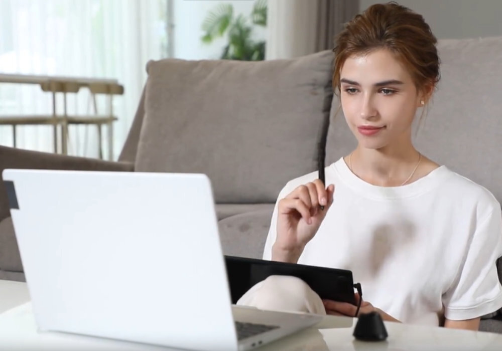 Osoba siedzi na kanapie, trzymając tablet graficzny i rysik, przed nią znajduje się otwarty laptop. W tle widoczny jest jasny salon z roślinami i stołem.
