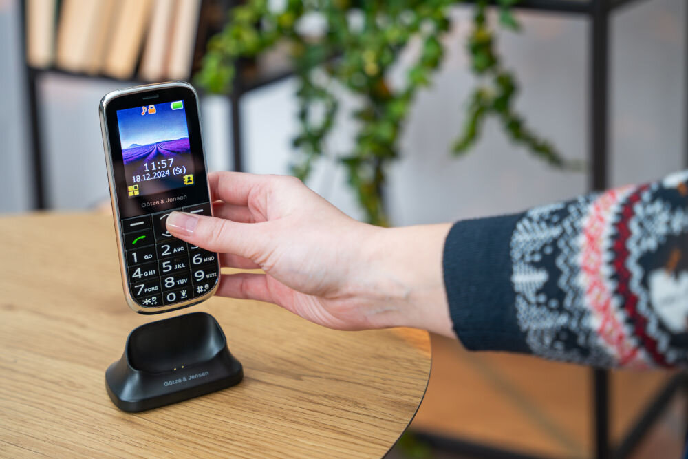 Osoba trzyma telefon z dużymi przyciskami i kolorowym wyświetlaczem, na którym widnieje godzina 11:57 i data 18.12.2024 (Śr). Telefon znajduje się na podstawce z napisem \'GÖTZE & JENSEN\'.