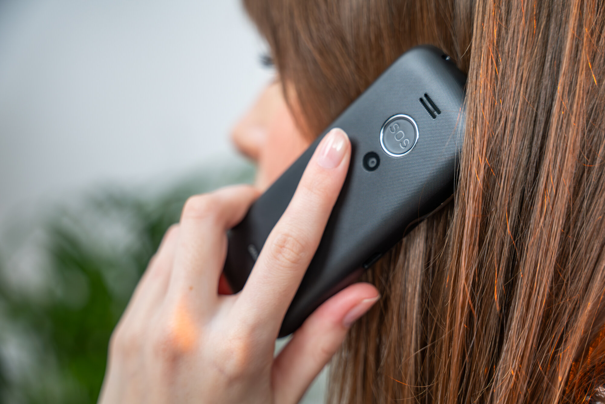 Osoba trzyma przy uchu czarny telefon z widocznym przyciskiem \'SOS\' na tylnej obudowie. W tle znajduje się rozmyta roślinność.