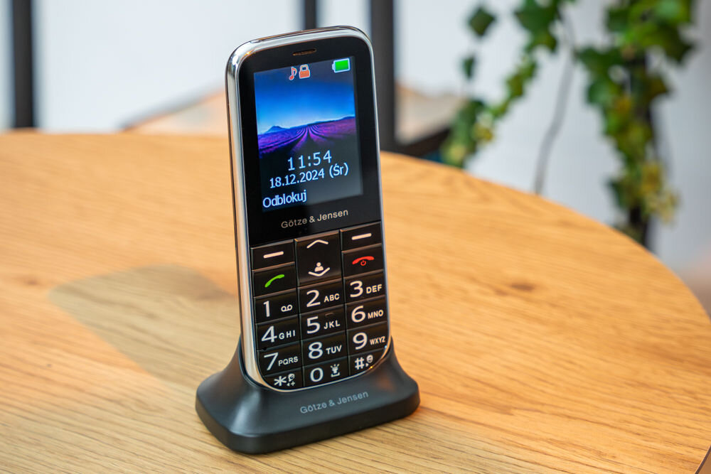 Telefon komórkowy Götze & Jensen GFE39 w kolorze czarnym stoi na podstawce na drewnianym stole. Na ekranie widoczna jest godzina 11:54, data 18.12.2024 (śr) oraz napis \'Odblokuj\'.