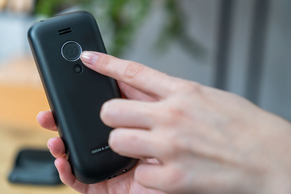 Osoba trzyma czarny telefon z widocznym przyciskiem SOS na tylnej obudowie. Na dole obudowy znajduje się napis \'GÖTZE & JENSEN\'.