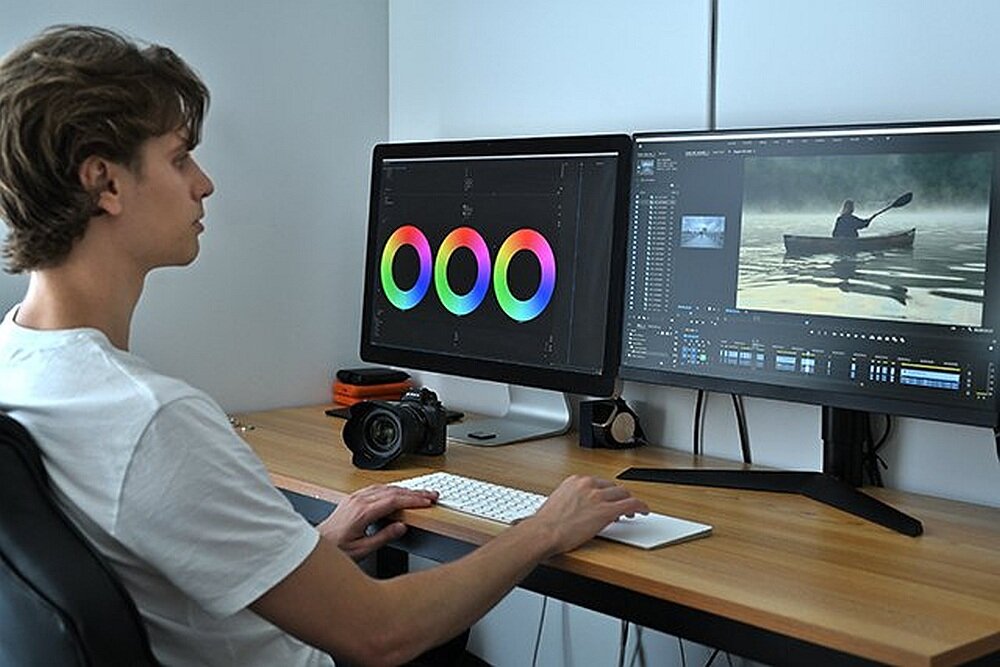 Osoba siedzi przy biurku z dwoma monitorami, na których widoczne są narzędzia do edycji wideo i obraz kajakarza na wodzie. Na biurku znajduje się aparat fotograficzny i klawiatura.