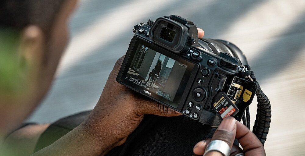 Osoba trzyma aparat bezlusterkowy marki Nikon, na ekranie którego widoczny jest podgląd fotografowanej sceny. Obok aparatu znajduje się karta pamięci o pojemności 128 GB.