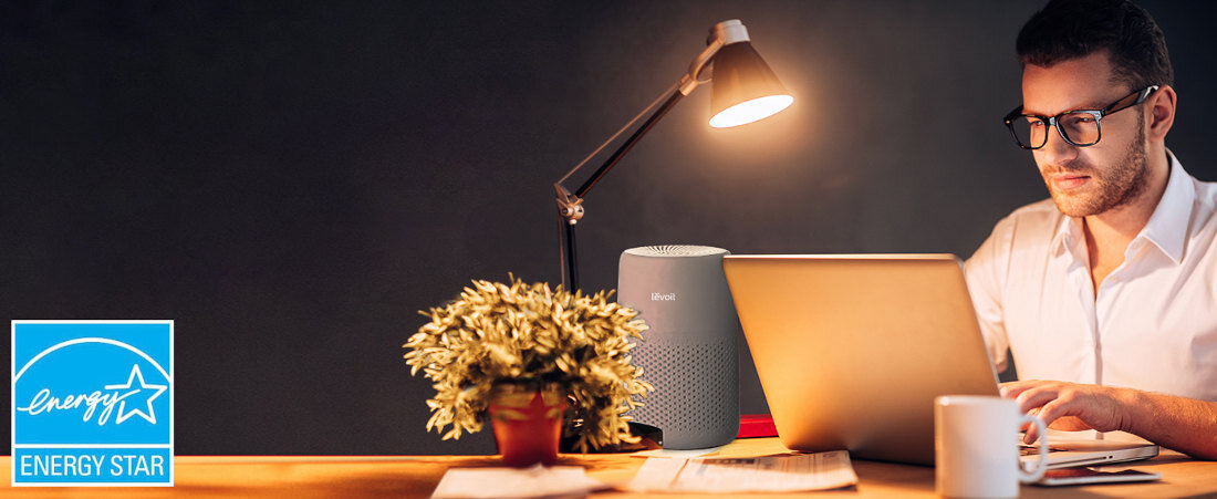 Na biurku znajduje się szary oczyszczacz powietrza LEVOIT Core Mini obok laptopa i lampki. W lewym dolnym rogu widoczne jest logo \'ENERGY STAR\'.