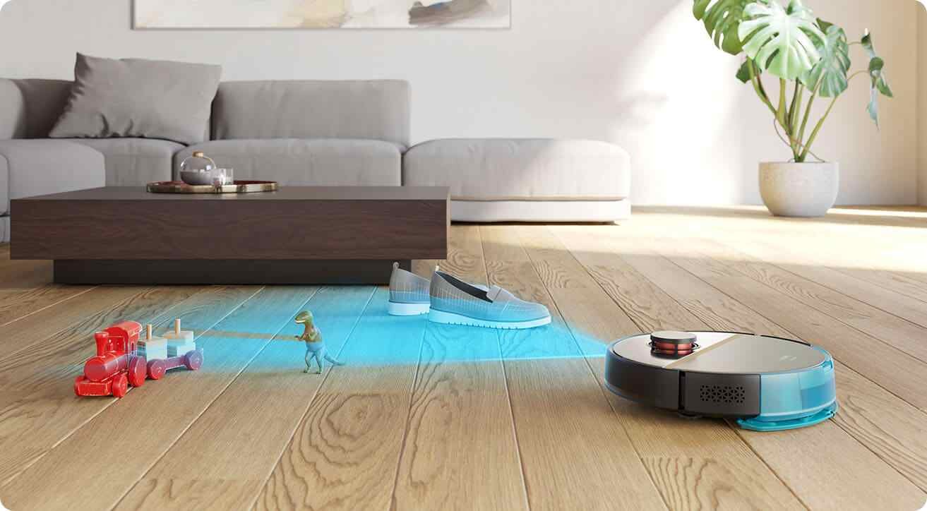 Robot sprzątający porusza się po drewnianej podłodze w salonie, omijając zabawki i buty. W tle widoczna jest szara sofa i stolik kawowy.