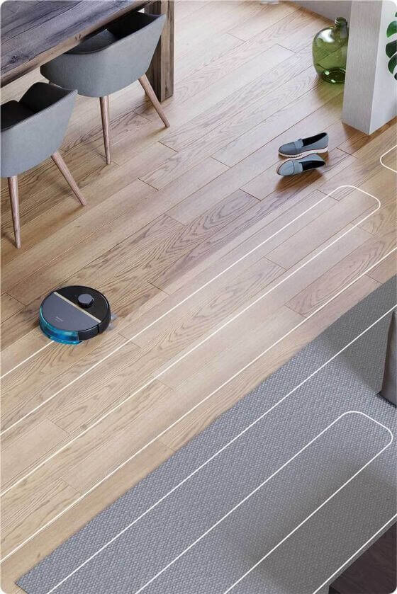 Na drewnianej podłodze widać robot sprzątający, obok którego leżą buty. W tle znajdują się krzesła przy stole oraz zielony wazon.