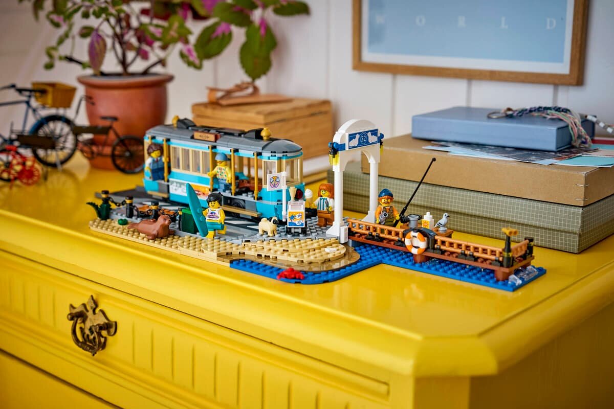 Na żółtej komodzie znajduje się zestaw LEGO przedstawiający tramwaj przy plaży z figurkami i akcesoriami. W tle widoczne są rośliny doniczkowe i pudełka.