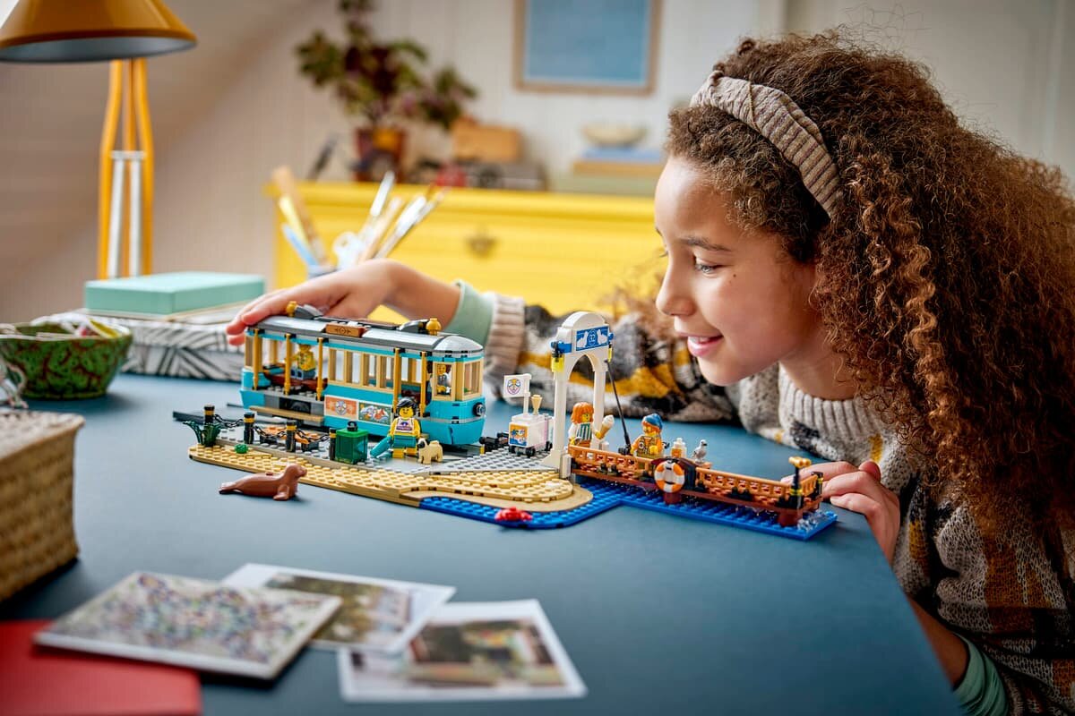 Na stole znajduje się zestaw LEGO przedstawiający tramwaj przy plaży z różnymi elementami, takimi jak figurki i budynki. W tle widać osobę bawiącą się zestawem.