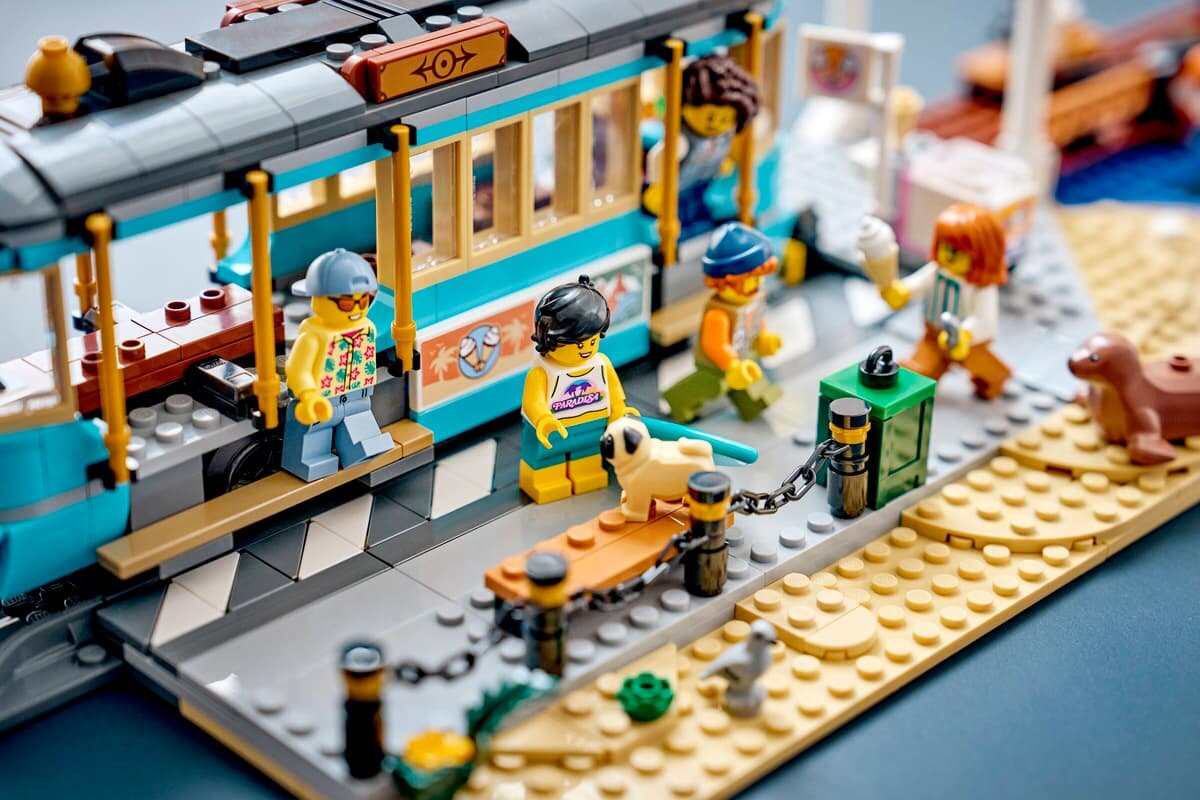 Zestaw LEGO przedstawiający tramwaj przy plaży z kilkoma figurkami postaci i psem. W tle widoczna jest plaża z piaskiem i elementami dekoracyjnymi.