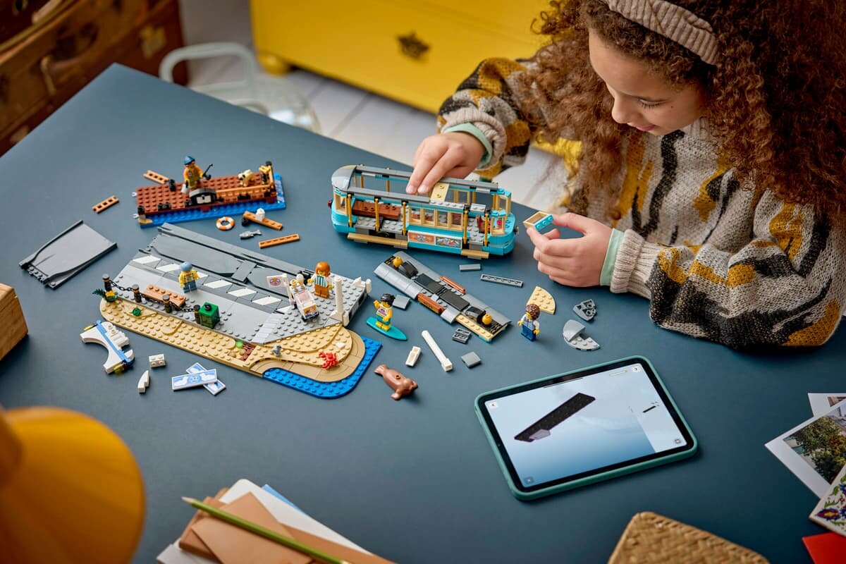 Na stole znajduje się zestaw klocków LEGO przedstawiający tramwaj i plażę, obok leży tablet z instrukcją. Osoba składająca zestaw jest widoczna w górnej części obrazu.
