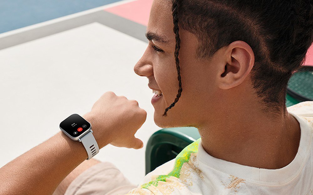 Osoba siedzi na zewnątrz, patrząc na smartwatch na nadgarstku z białym paskiem i czarnym ekranem. Na ekranie widoczne są ikony połączenia i wiadomości.