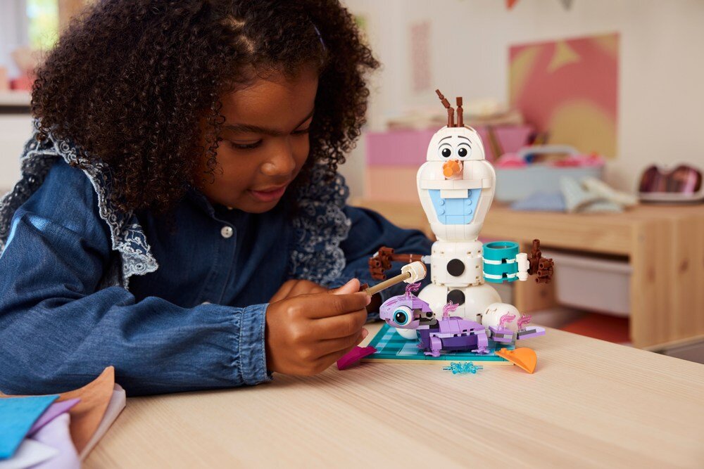 Dziecko bawi się zestawem LEGO przedstawiającym postać Olafa z filmu animowanego, z dodatkowymi elementami na stole. W tle widać rozmyte elementy pokoju dziecięcego.