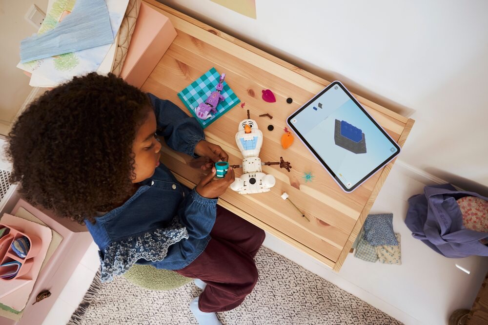 Dziecko bawi się zestawem LEGO przedstawiającym postać Olafa na drewnianym stole, obok leży tablet z instrukcją budowy. Na stole znajdują się również różne elementy LEGO i kolorowe tkaniny.