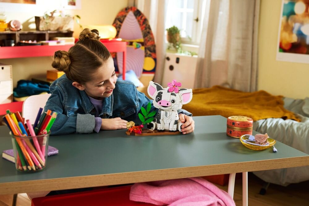Dziecko siedzi przy stole i bawi się zestawem LEGO przedstawiającym postać z dużymi uszami i różowym kwiatkiem na głowie. Na stole znajdują się również kolorowe kredki w szklanym pojemniku oraz kilka innych przedmiotów.