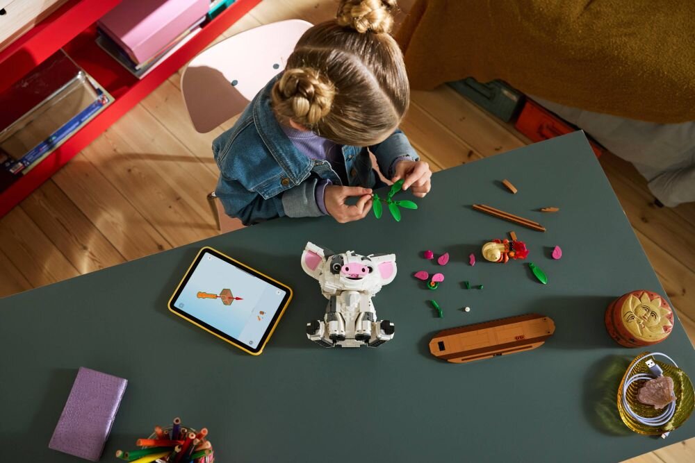 Dziecko siedzi przy stole, bawiąc się zestawem LEGO z figurką przypominającą małą świnkę. Obok leży tablet z wyświetlonym obrazem oraz różne elementy LEGO.