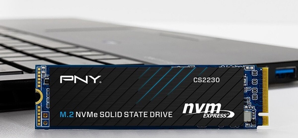 Na pierwszym planie znajduje się dysk SSD z oznaczeniami: PNY, CS2230, M.2 NVMe SOLID STATE DRIVE, nvm express. W tle widoczny jest fragment otwartego laptopa.
