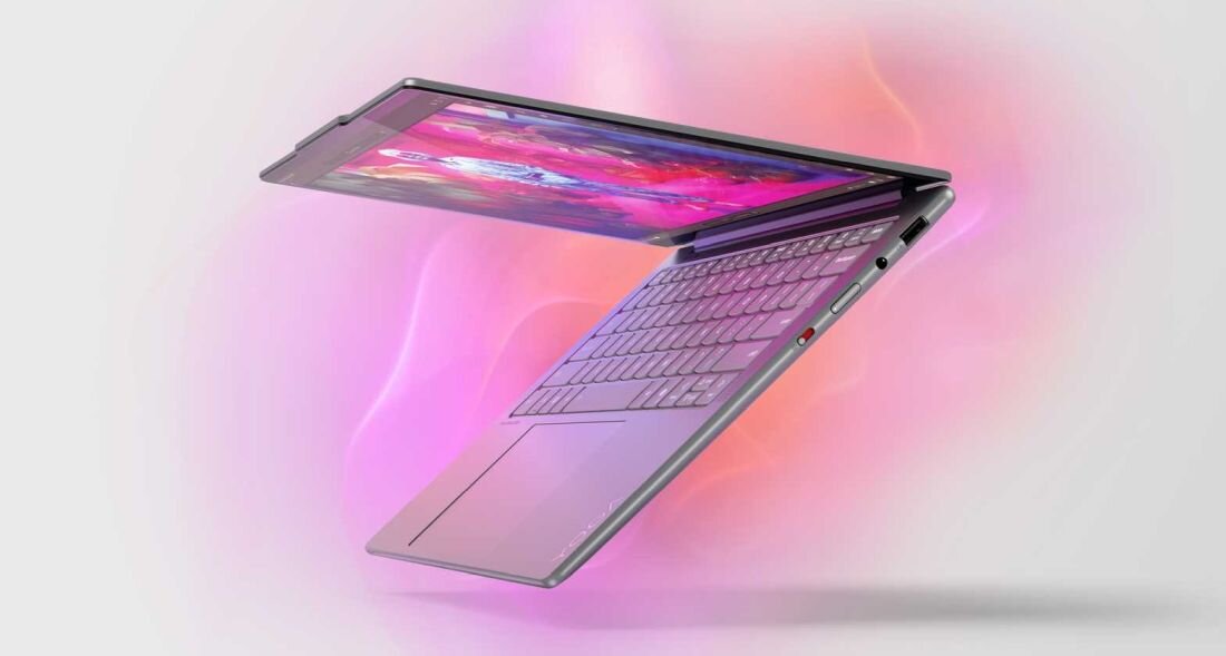 Laptop w kolorze srebrnym jest otwarty pod kątem, ukazując klawiaturę i ekran z kolorowym wyświetlaczem. Tło jest rozmyte w odcieniach różu i fioletu.