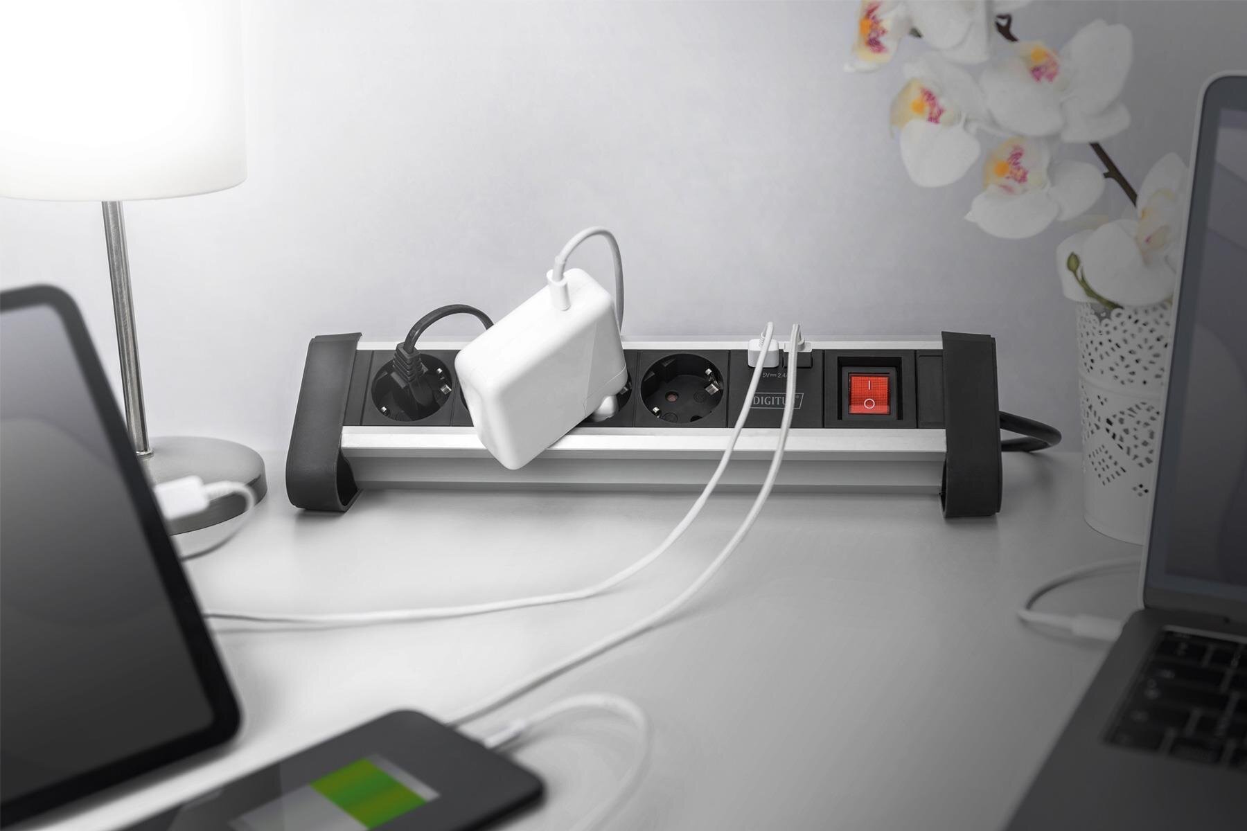 Na biurku znajduje się aluminiowa listwa zasilająca z czterema gniazdami i dwoma portami USB, do której podłączone są różne urządzenia. Obok listwy stoi lampa i doniczka z białymi kwiatami.