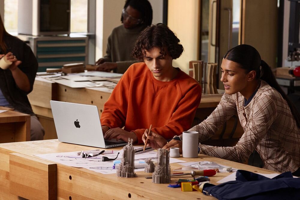 APPLE MacBook Pro 2026 16 Retina M5 Pro 1TB SSD macOS 12 MP Center Stage dostosowując ujęcie do jego ruchów Desk View przestrzeni roboczej - Dwie osoby pracujące przy drewnianym stole w warsztacie, korzystające z otwartego urządzenia i notatek