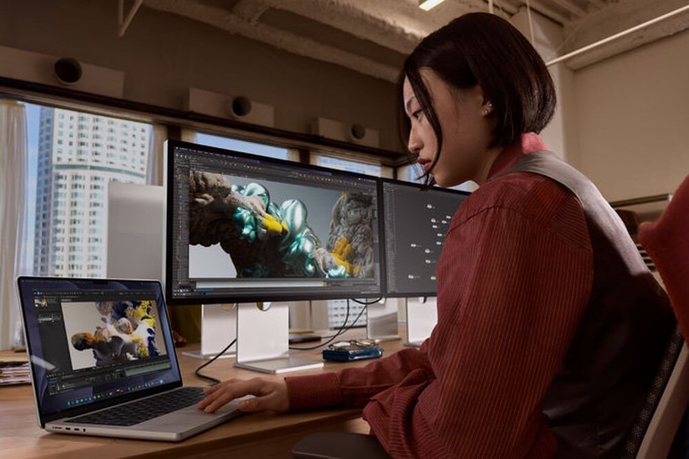 APPLE MacBook Pro 2026 16 Retina M5 Pro 1TB SSD macOS dwóch zewnętrznych wyświetlaczy ekran laptopa dodatkowe monitory - Kobieta pracująca przy biurku z otwartym urządzeniem mobilnym oraz dwoma monitorami wyświetlającymi grafikę 3D