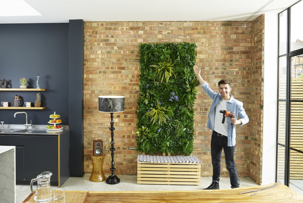 Wiertarka udarowa BLACK&DECKER BEH710-QS pomieszczenie osoba stoi przed ścianą z wiertarką w dłoni wskazuje dłonią na ścianę na ścianie założona ścianka z roślin funkcjonalność wygodne użytkowanie wiercenie otworów o średnicy do 25 mm w drewnie do 13 mm w betonie do 10 mm w stali wysoka precyzja działania mocny udar