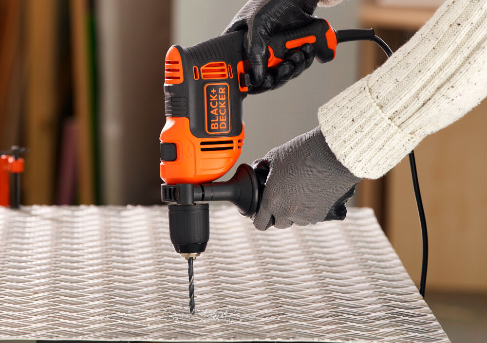 Wiertarka udarowa BLACK&DECKER BEH710-QS pomieszczenie metalowy blat osoba trzyma wiertarkę w dłoni wierci otwór w metalowym blacie uchwyt pomocniczy zwiększa stabilność prowadzenia poprawia bezpieczeństwo pracy główna rękojeść ma ergonomiczną konstrukcję ułatwia prowadzenie narzędzia ogranicza zmęczenie dłoni antypoślizgowa powłoka lepsza kontrola nad wiertarką