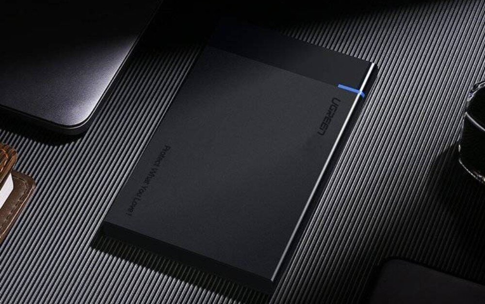 Obudowa dysku UGREEN 30847 Czarna, prostokątna obudowa dysku leżąca na prążkowanej powierzchni. Na obudowie widoczne jest logo Ugreen, mała niebieska dioda oraz pionowy napis