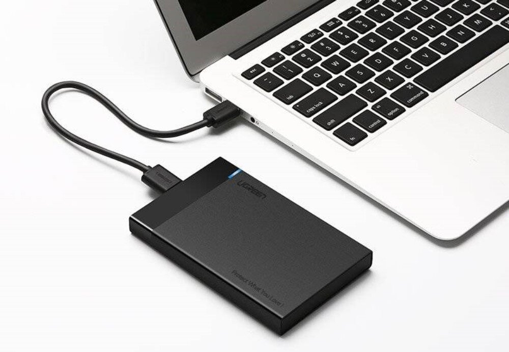 Obudowa dysku UGREEN 30847 Obudowa dysku połączona czarnym kablem z portem USB w srebrnym laptopie. Urządzenie spoczywa na białym blacie obok klawiatury
