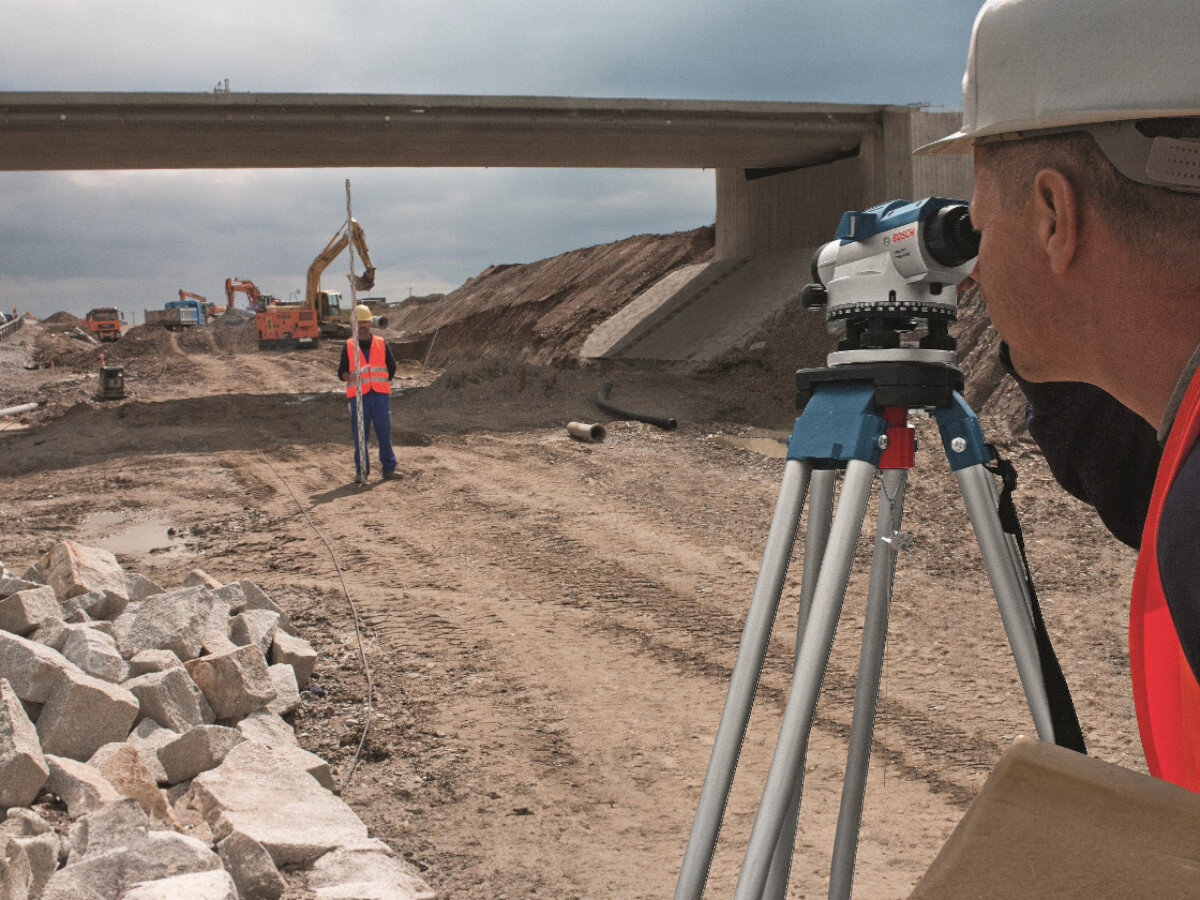 Osoba w kasku i kamizelce odblaskowej używa niwelatora BOSCH na statywie na placu budowy. W tle widoczne są maszyny budowlane i most.