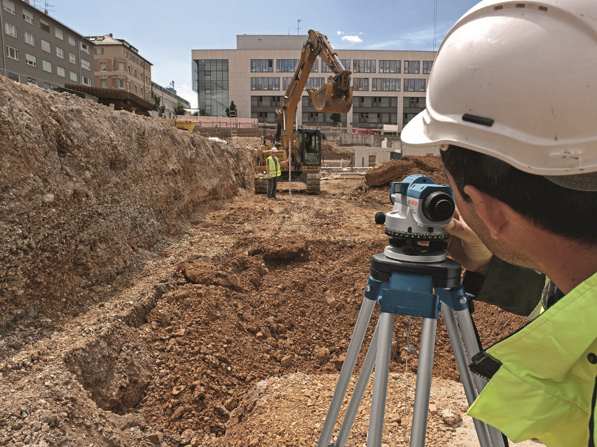 Osoba w kasku obsługuje niwelator na statywie na placu budowy. W tle widoczna jest koparka oraz budynki.