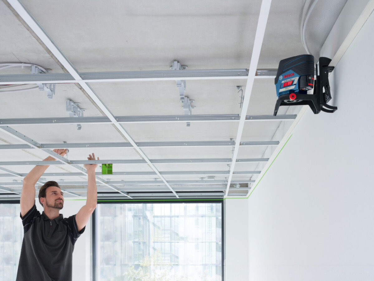 Osoba pracuje przy suficie podwieszanym, używając lasera liniowego zamontowanego na ścianie. Na urządzeniu widoczne jest logo \'BOSCH\'.
