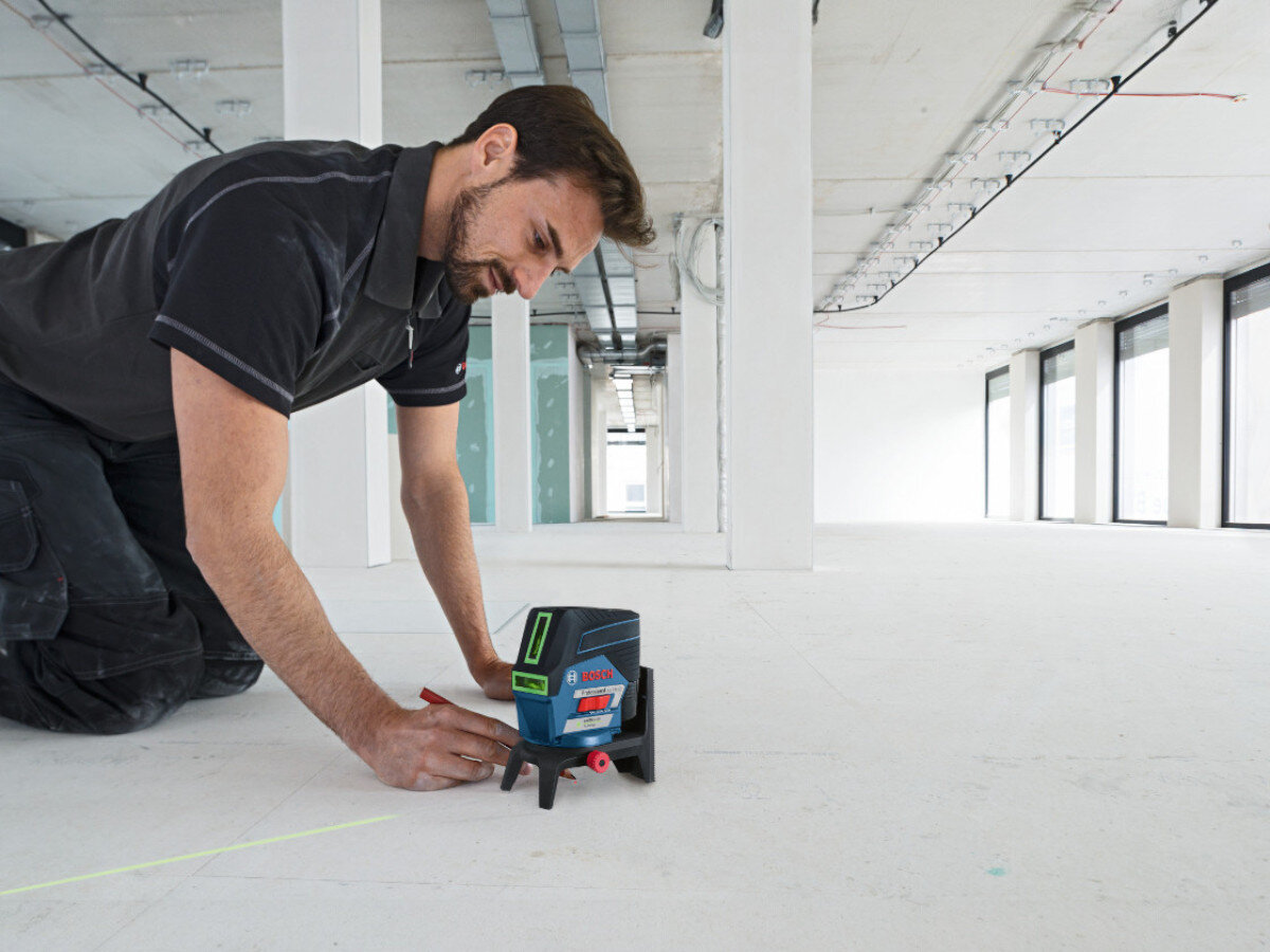 Osoba klęczy na podłodze w jasnym, pustym pomieszczeniu, używając lasera liniowego BOSCH GCL 2-50 CG. Urządzenie emituje zieloną linię na podłodze.