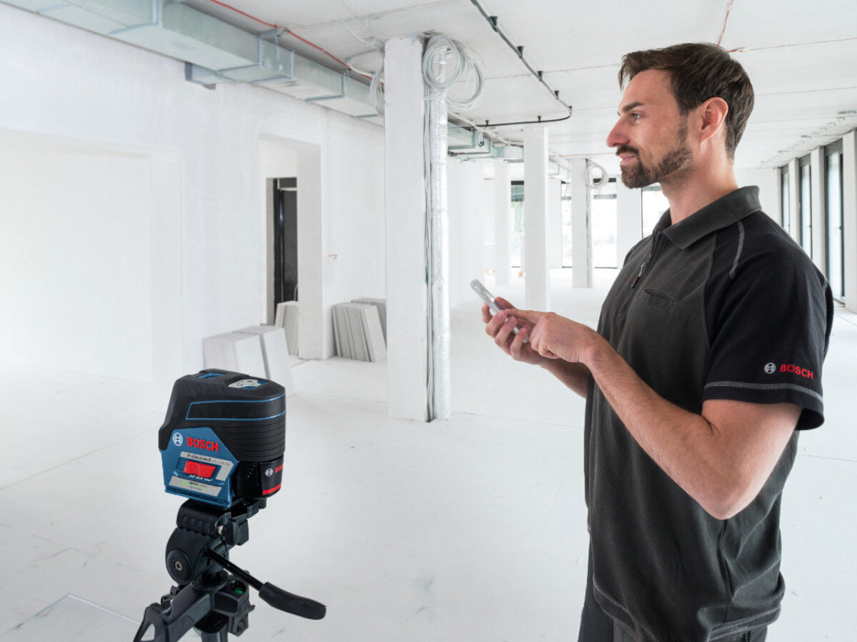 Mężczyzna w ciemnej koszulce stoi w jasnym pomieszczeniu, trzymając w ręku urządzenie. Na statywie znajduje się laser liniowy z widocznym logo BOSCH.