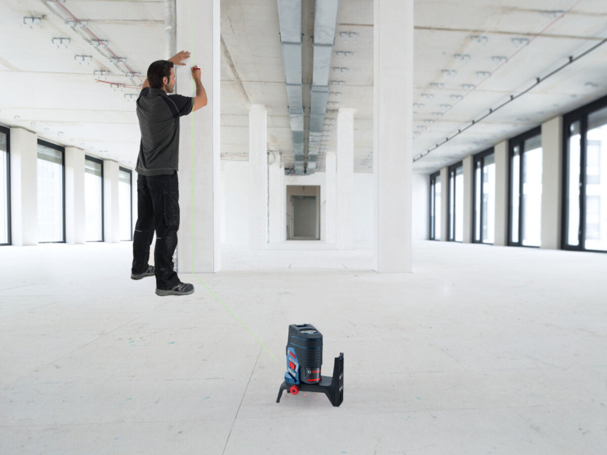 W dużym, jasnym pomieszczeniu znajduje się laser liniowy ustawiony na podłodze, emitujący zieloną linię. W tle osoba pracuje przy ścianie, używając narzędzia pomiarowego.