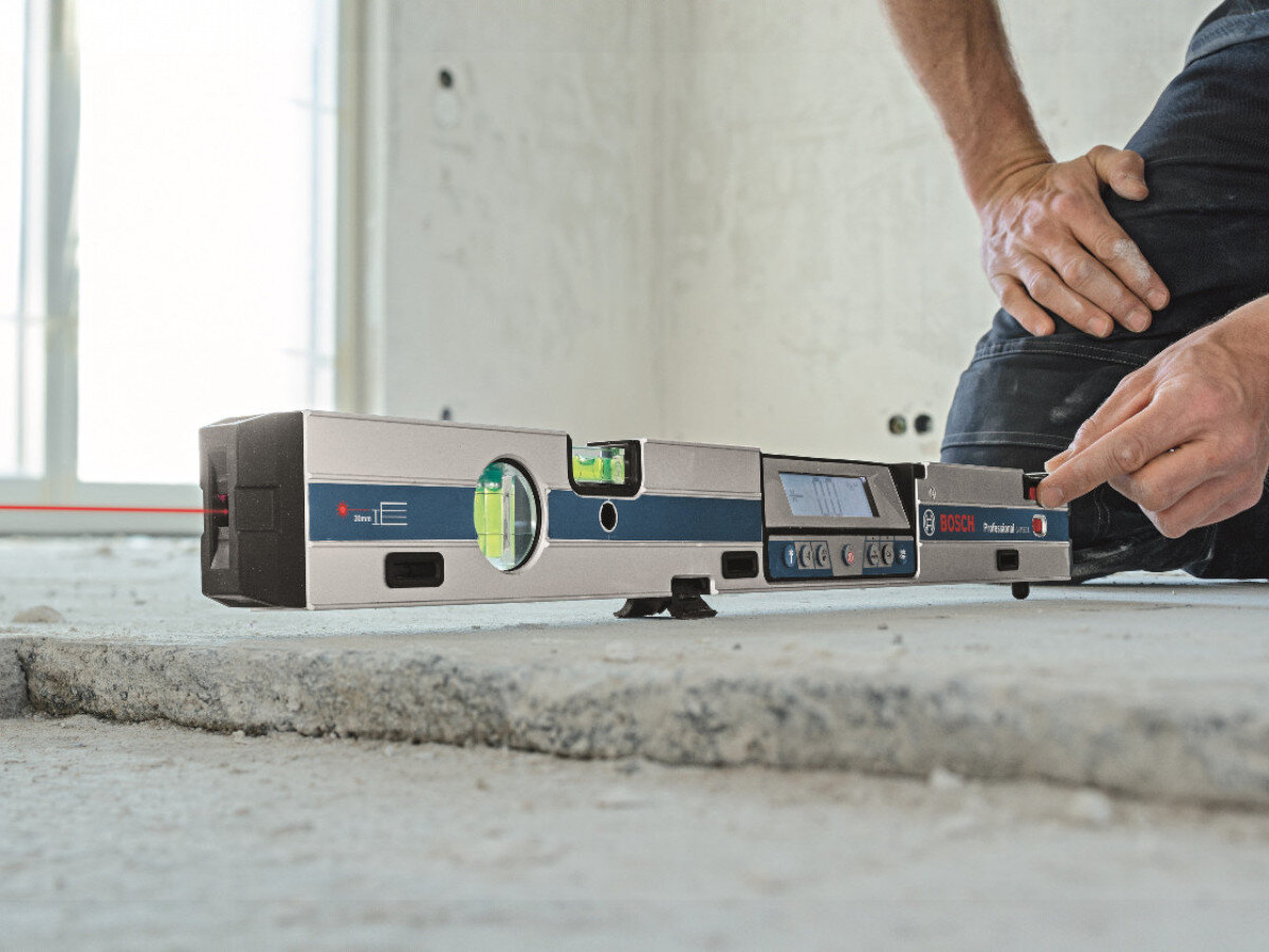 Poziomnica laserowa BOSCH GIM 60 L Professional leży na betonowej powierzchni, a obok niej znajduje się osoba w ciemnym ubraniu. Na poziomnicy widoczne jest logo BOSCH oraz kilka przycisków i wyświetlacz.
