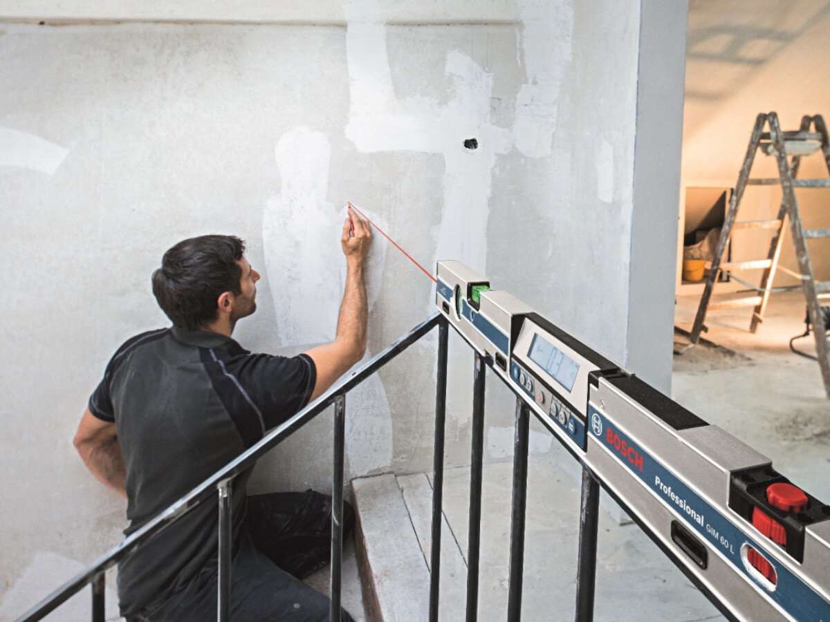 Osoba używa poziomnicy laserowej BOSCH GIM 60 L Professional do wyznaczania linii na ścianie. W tle widoczna jest drabina i jasne pomieszczenie w trakcie remontu.
