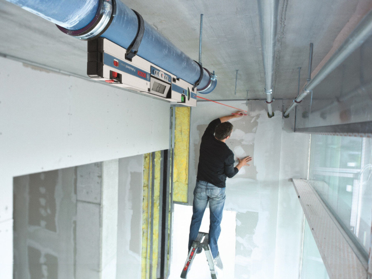 Osoba stoi na drabinie i pracuje przy ścianie w pomieszczeniu. Na suficie zamontowana jest poziomnica laserowa z widocznym logo BOSCH.