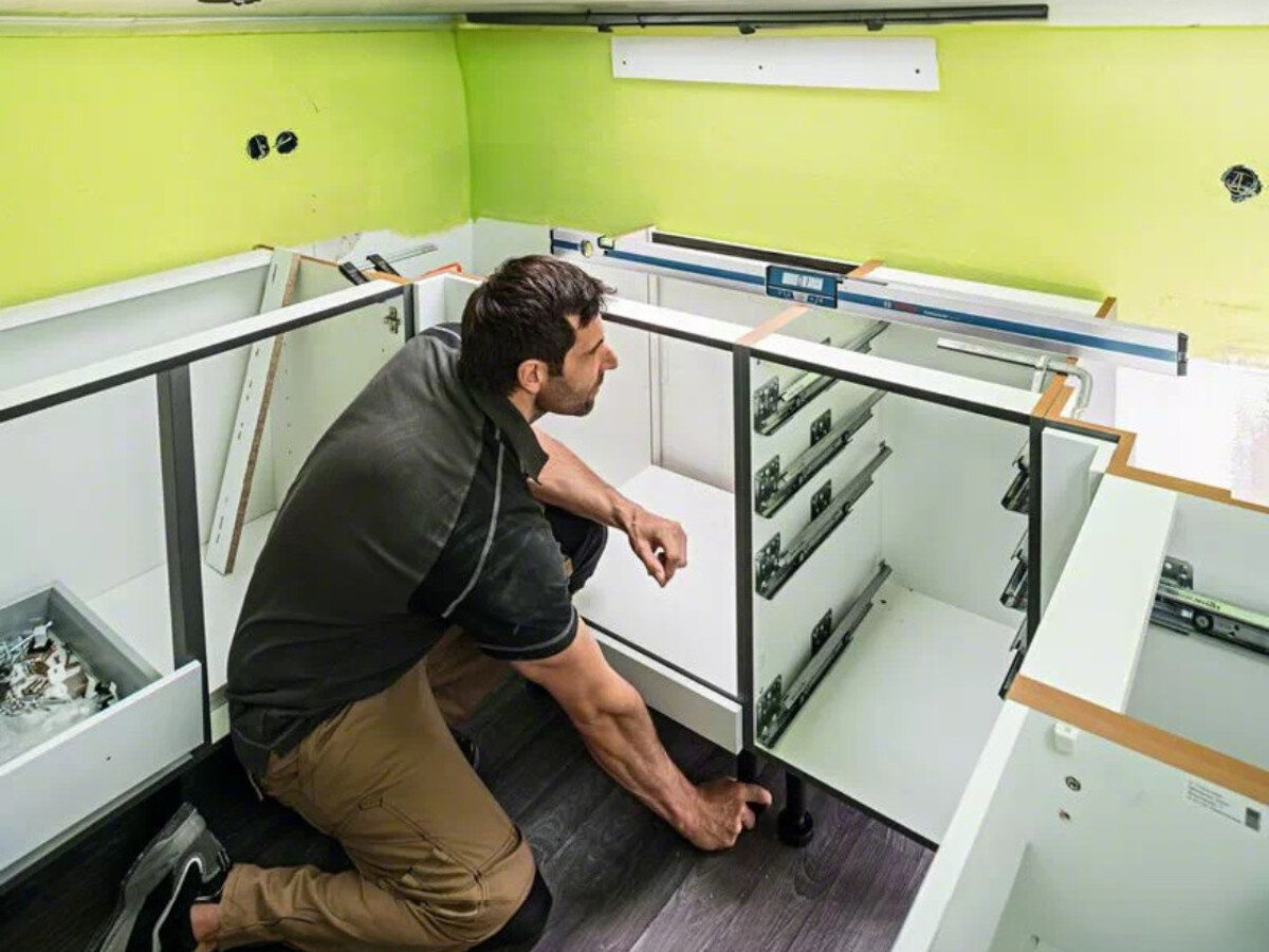 Osoba montuje szafki kuchenne w pomieszczeniu o jasnozielonych ścianach. Na blacie znajduje się poziomnica elektroniczna z widocznym logo BOSCH.