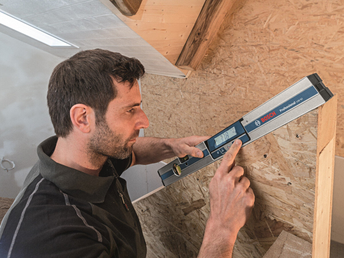 Osoba używa elektronicznej poziomnicy marki BOSCH, trzymając ją przy drewnianej powierzchni. Na poziomnicy widoczny jest wyświetlacz z cyframi oraz logo BOSCH Professional.