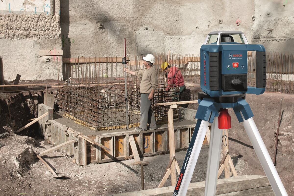 Na placu budowy znajduje się laser rotacyjny BOSCH Professional ustawiony na statywie. W tle widać dwóch pracowników przy zbrojeniu fundamentów.