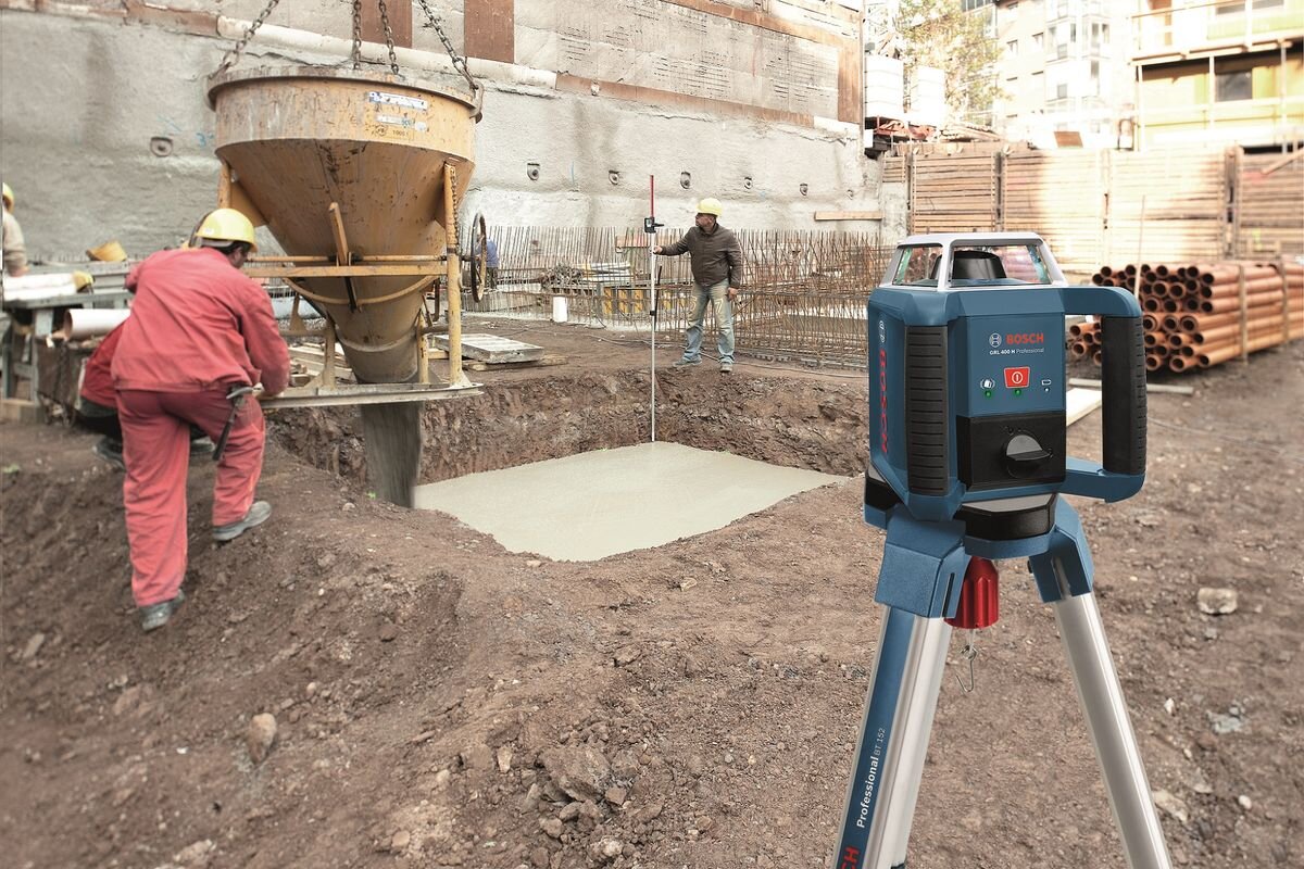 Na placu budowy widać laser rotacyjny BOSCH Professional GRL 400 H ustawiony na statywie. W tle pracownicy w kaskach pracują przy wylewaniu betonu do wykopu.
