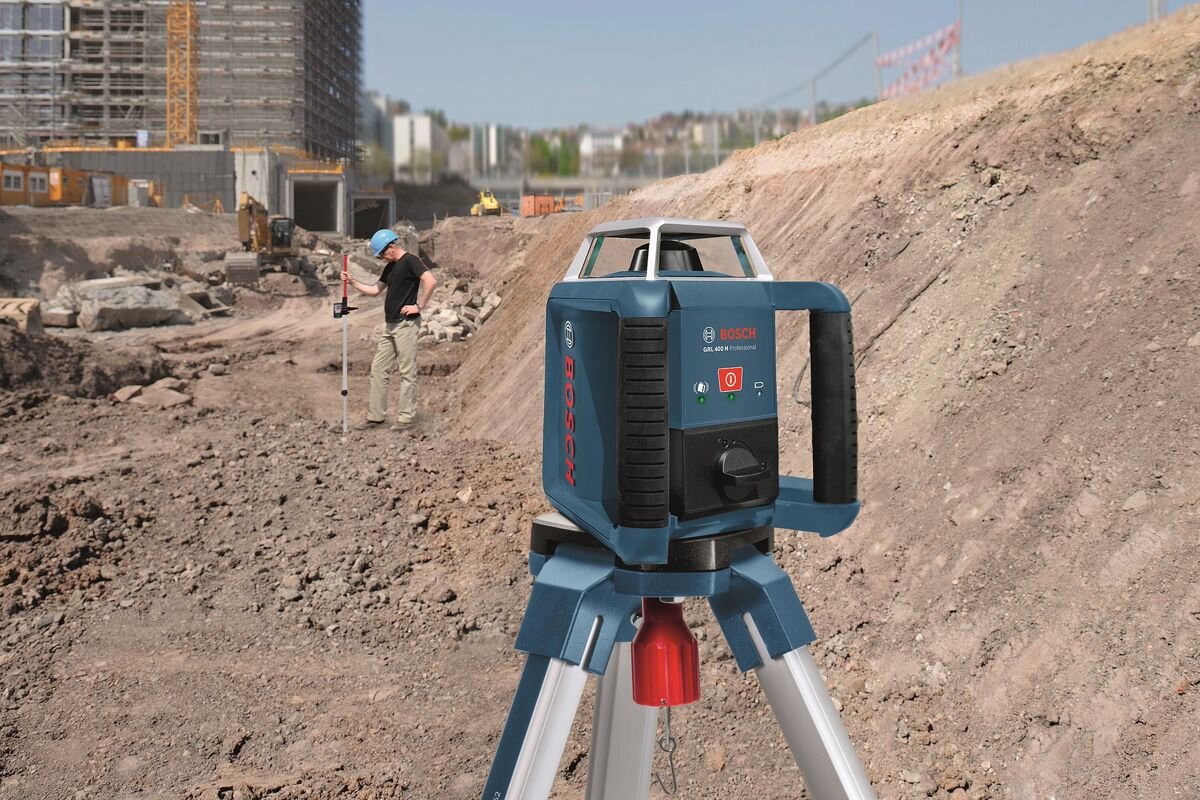 Na placu budowy znajduje się laser rotacyjny BOSCH Professional GRL 400 H umieszczony na statywie. W tle widoczny jest pracownik z narzędziem pomiarowym oraz budynki i sprzęt budowlany.