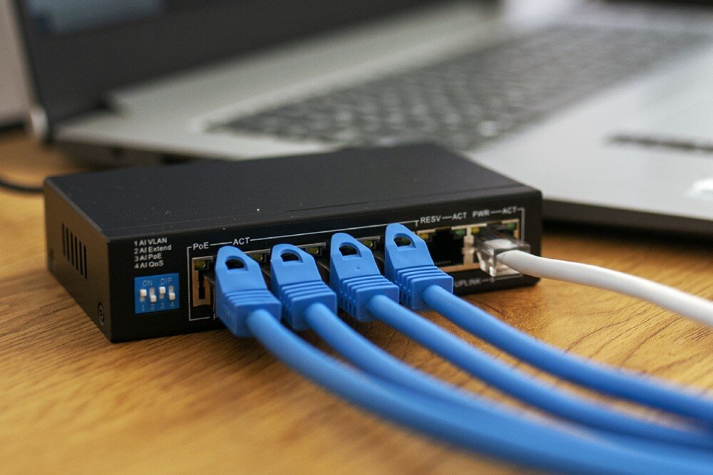 Switch EXTRALINK Euros V2 EX.14831 Switch sieciowy na drewnianym blacie, do czterech portów wpięte niebieskie kable ethernetowe, jeden biały kabel połączony z komputerem przenośnym