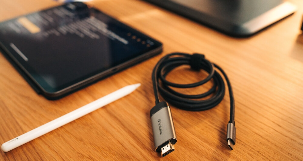 Na drewnianym stole leży zwinięty kabel z końcówkami USB Typ-C i HDMI, obok którego znajduje się tablet oraz biały rysik. Na obudowie kabla widoczne jest logo \'Verbatim\'.