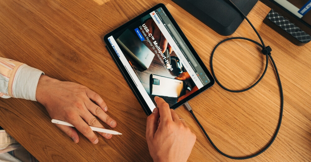 Tablet na drewnianym stole z wyświetloną stroną internetową, do którego podłączony jest kabel USB-C. Obok leży dłoń trzymająca rysik.