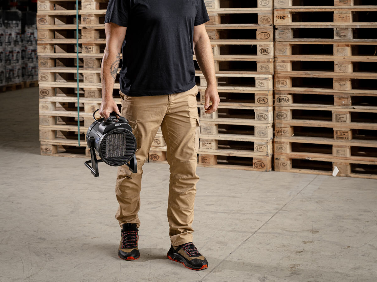 Nagrzewnica elektryczna DEDRA DED9931C2, uchwyt, metal, ergonomia, trwałość, Mężczyzna w beżowych spodniach niesie nagrzewnicę DEDRA DED9931C2 w magazynie z paletami.